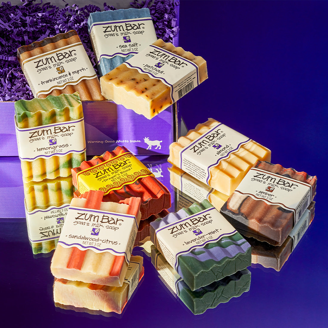 Almond, Amber, Dragon's Blood, Frankincense & Myrrh, Lavender-Mint, Lemongrass, Patchouli, Sandalwood-Citrus, and Sea Salt labeled wavy soap bars inside spilling out of purple gift box. Purple to blue gradient background.