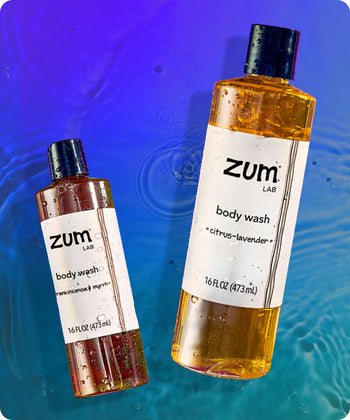 Frankincense & Myrrh and Citrus-Lavender scented bottles of body wash flying in the air with water droplets on them. Watery Blue to green gradient background.