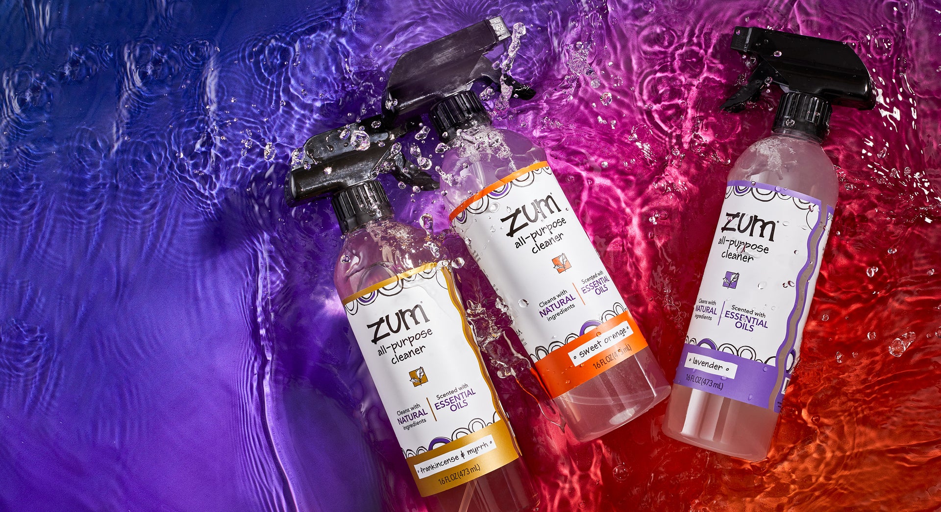 Frankincense & Myrrh, Sweet Orange, and Lavender scented bottles with sprayers of All-Purpose Cleaner. The three bottles are laying in a bed of water with splashes all around. Blue to purple to orange gradient background.