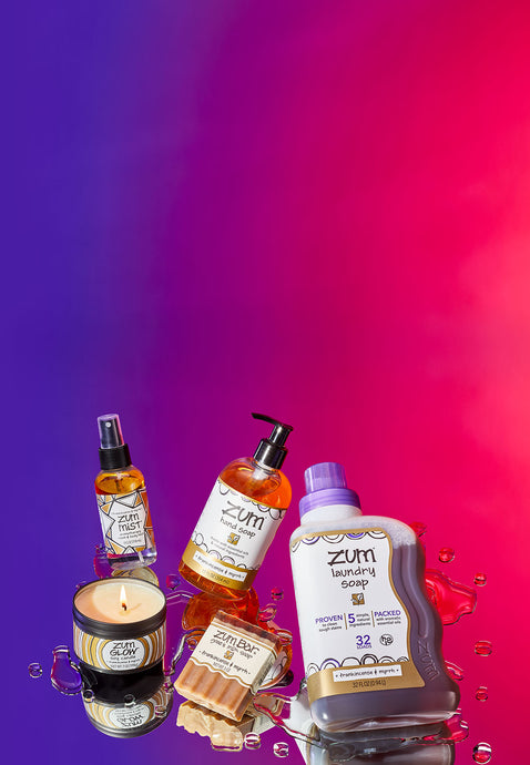 Frankincense & Myrrh grouping of products comprised of a soy candle in a tin, a mist bottle, a hand soap bottle, a waxy bar soap, and a 32 fl oz laundry soap bottle. Products are laying at an angle on a mirrored surface with a purple to red background.