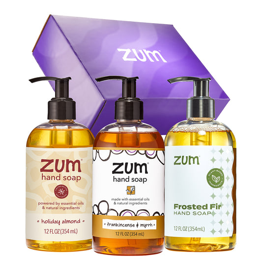 A three-bottle hand soap gift set from Zum with a purple box behind it. The set includes Holiday Almond, Frankincense & Myrrh, and Frosted Fir hand soaps in clear pump bottles filled with amber liquid, shown on a white background.