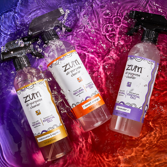 Frankincense & Myrrh, Sweet Orange, and Lavender scented sprayed bottles laying down splashed with water on a purple to orange gradient background.
