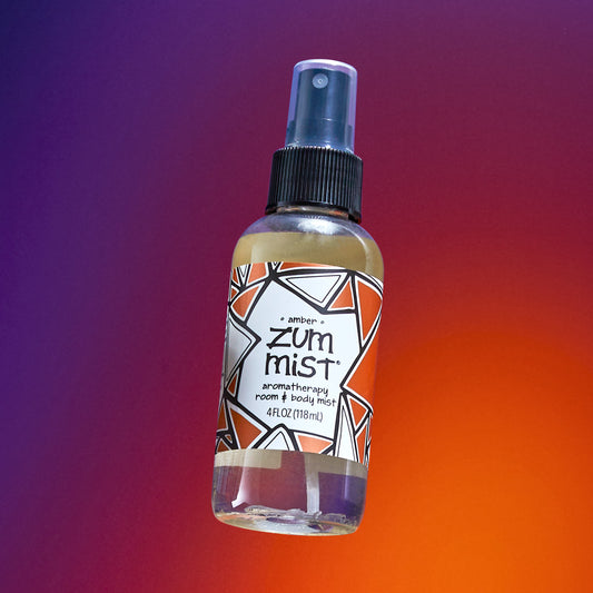 Amber scented room & body spray mist bottle flying in the air with a purple to orange gradient background.