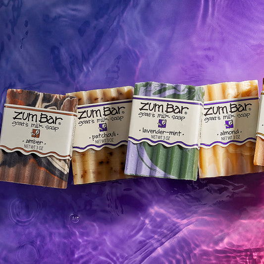 Amber, Patchouli, Lavender-Mint, and Almond scented bar soaps on a watered surface. Purple to pink gradient background.