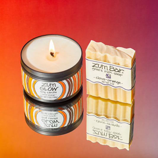 Clove-orange scented soy candle in a tin and wavy bar soap on a mirrored surface with a orange to pink gradient in the background.