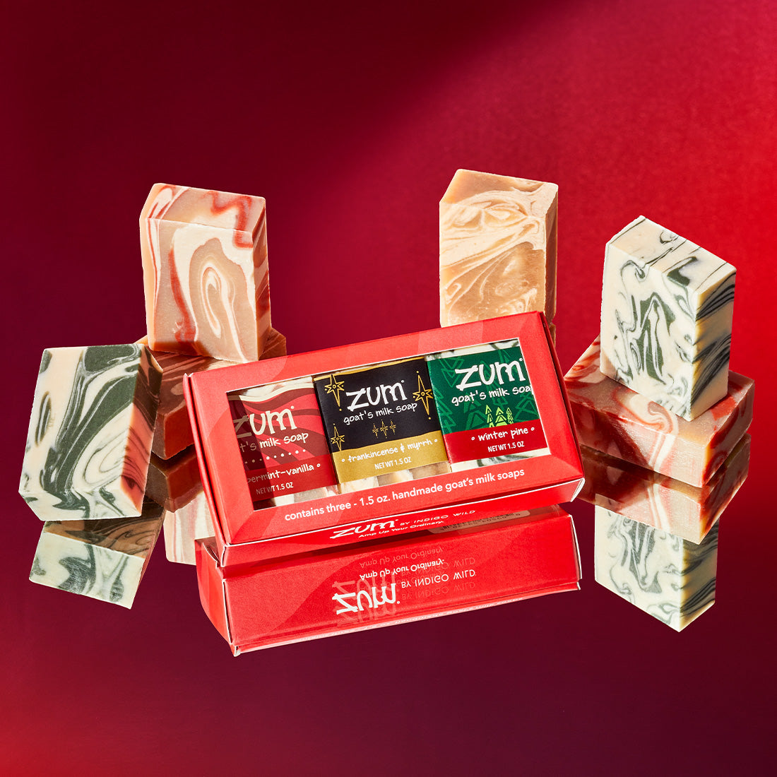Fold in red box with rectangular cut out in front that hold Peppermint-Vanilla, Frankincense & Myrrh, and Winter Pine mini bar soaps. Other unlabeled bar soaps with swirls of the same scents as the ones in the box are stacked and surrounding the box on a mirrored surface. Red gradient background.