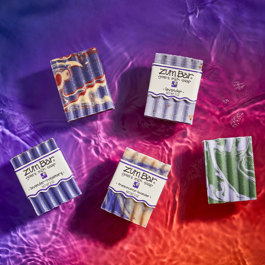 Lavender-Rosemary, Lavender-Lemon & Patchouli, Frankincense-Lavender, Lavender, and Lavender-Mint scented waxy bar soaps laying on a watered surface. Purple to orange gradient background.