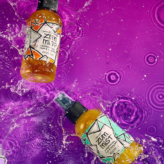 Patchouli-Orange and Sea Salt room & body spray bottles splashing on a purple watered surface.