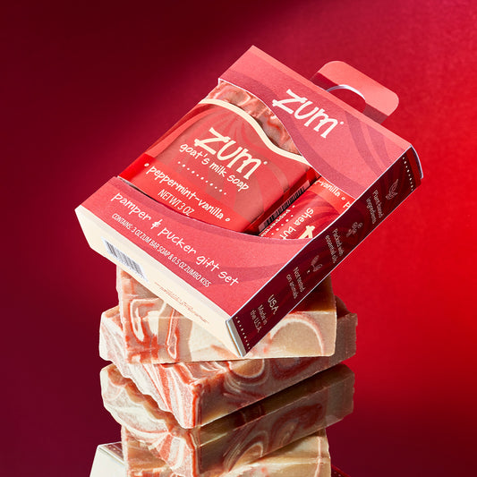 Pamper & Pucker red gift box wit ha wavy cutout in the middle filled with Peppermint-Vanilla scented bar soap and 0.5 oz lip balm. This box is sitting on stacked unlabeled Peppermint-Vanilla scented bar soaps at an angle. Mirrored surface underneath with a red gradient background.