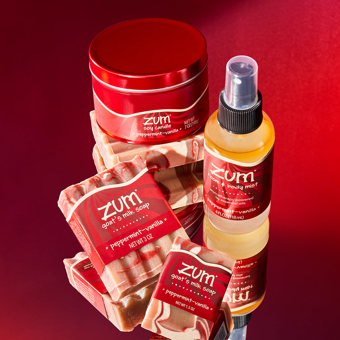 Peppermint-Vanilla scented product grouping sitting on mirrored surface with a red gradient background. Mini bar soap in the front, a bar soap behind it to the left, to its right a mist bottle and behind them a soy candle in a red tin sitting on a stack of unlabeled bar soaps.