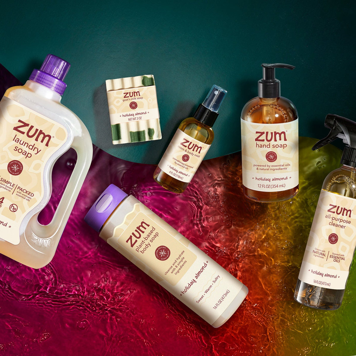 A set of Zum products arranged on a glossy, multicolored water surface against a dark teal background. The products include a large bottle of Holiday Almond laundry soap, a hand soap bottle, an all-purpose cleaner, a liquid soap bottle, and a bar soap.
