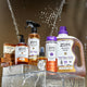 Assorted Zum products on acrylic boxes with water cascading behind and a brown background. Product left to right: stack of three Frnakincense & Myrrh Bar Soaps, Amber Hand Soap, Frankincense & Myrrh All-Purpose Cleaner, Frankincense & Myrrh Mist, Amber Body Soap, Frankincense & Myrrh Laundry Soap.