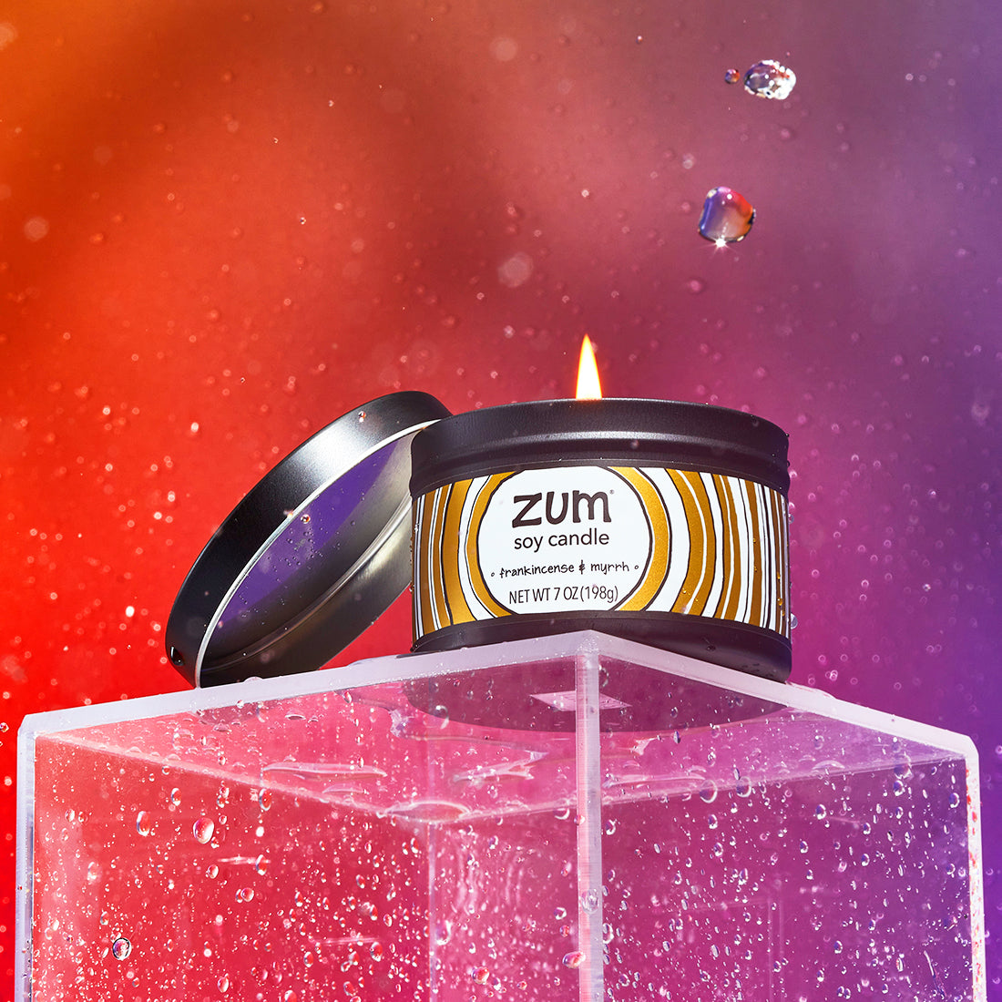 A lit Frankincense & Myrrh soy candle in a black tin with its lid resting against it. Candle is on a acrylic stand with water droplets falling around and an orange to purple gradient background.