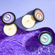 Top shot of three Zum soy candles, two lit with their lid off to the side in the scents Lavender and Amber, and a closed Sea Salt soy candle tin to the right. Amber is on a light purple wave, and Lavender and Sea Salt are on purple water.