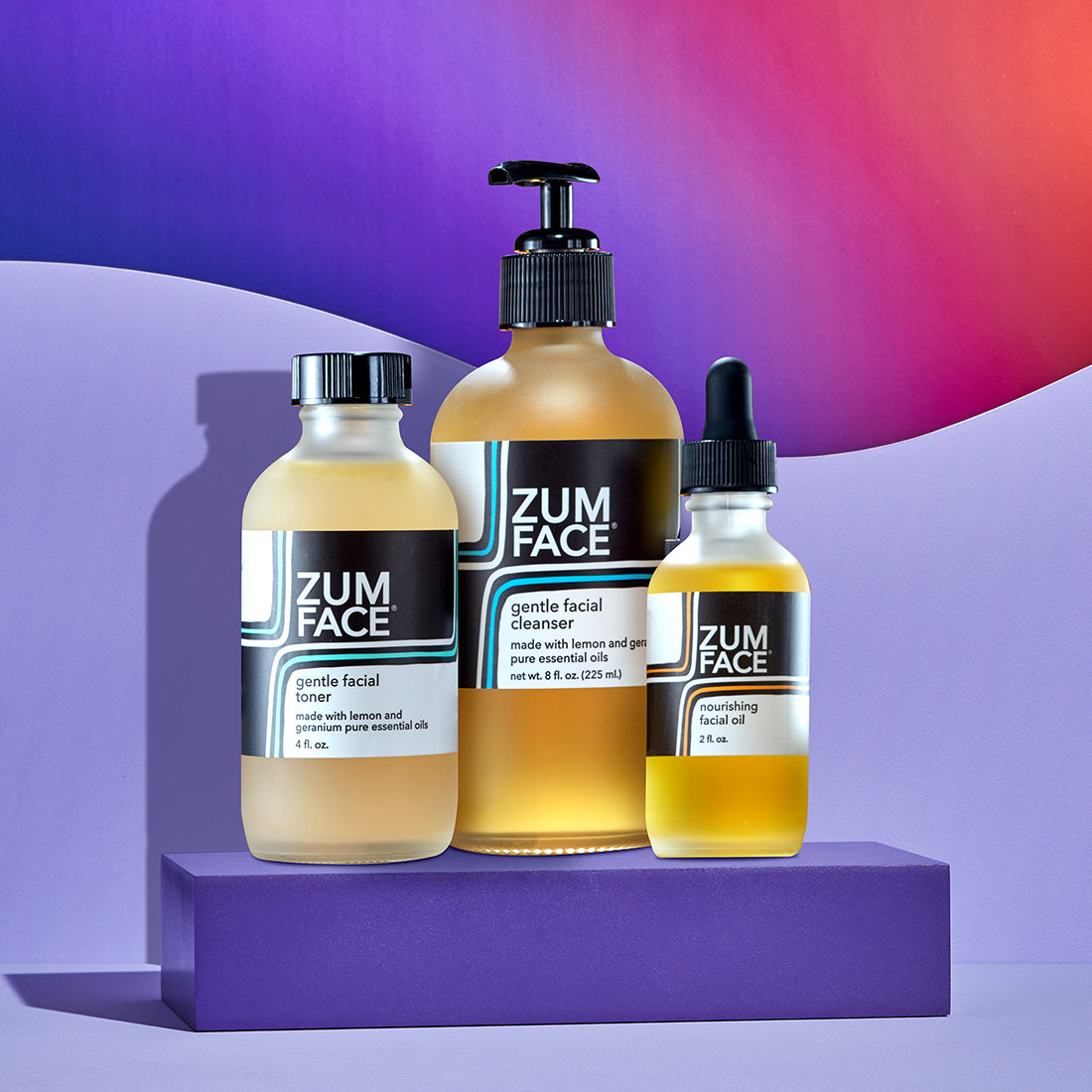 Zum Gentle Facial Toner, Gentle Facial Cleanser, and Nourishing Facial Oil bottles on a purple block. A light purple surface with a light purple wave behind the product and a purple to orange gradient background.
