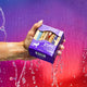 A hand coming from the left side of the image holding a 9 oz Bundle Box filled with slides of goat's milk soap. Water falling behind with a purple to pink gradient background.