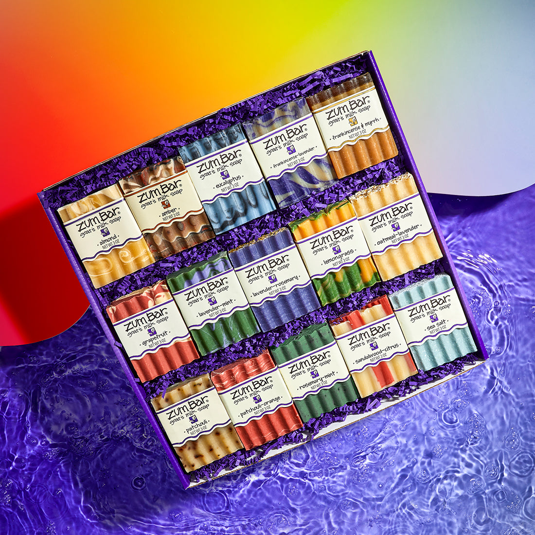 Purple gift box with 15 Bar Soaps in the scents Almond, Amber, Eucalyptus, Frankincense-Lavender, Frankincense & Myrrh, Grapefruit, Lavender-Mint, Lavender-Rosemary, Lemongrass, Oatmeal-Lavender, Patchouli, Patchouli-ORange, Rosemary-Mint, Sandalwood-Citrus, and Sea Salt. The box is half on a red to light purple gradient wavy surface and another half on a purple water.