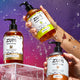 On the left side of the image is a Frankincense & Myrrh Hand Soap bottle with a pump on an acrylic stand, with two hands coming from the right side of the image holding a Lemongrass Hand Soap at the top and an Amber Hand Soap at the bottom. Water falling around and on the product. Blue, purple, pink gradient background.