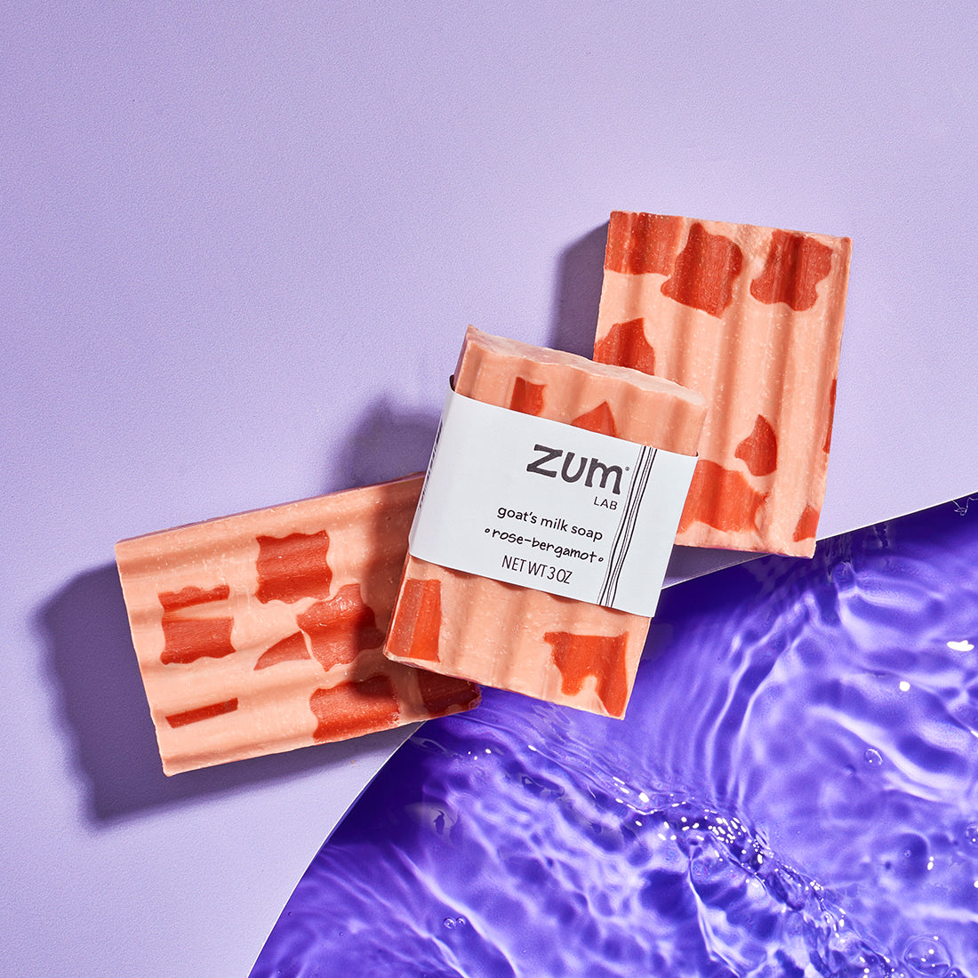 Two unlabeled Rose-Bergamot bar soaps with a labeled version on top, laying on a light purple wavy shape with purple water underneath.