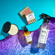 Tomato Leaf products half on a blue wavy shape, half on purple to blue water underneath. From left to right: Tomato Leaf Soy Candle, Room & Body Spray, Goat's Milk Soap, and All-Purpose Cleaner.