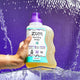 A hand on a Sea Salt 32 fl oz Laundry Soap on a clear acrylic box with water falling behind it with a purple background.