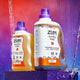 Sweet Orange 32 fl oz Laundry Soap and an Amber 64 fl oz Laundry Soap on a purple box. Water cascading behind and a orange, blue, and purple gradient background.
