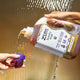 A hand coming in from the right holding a 64 fl oz Laundry Soap, pouring soap into a purple cap being held by a hand coming from the left. Water falling behind with a brown background.