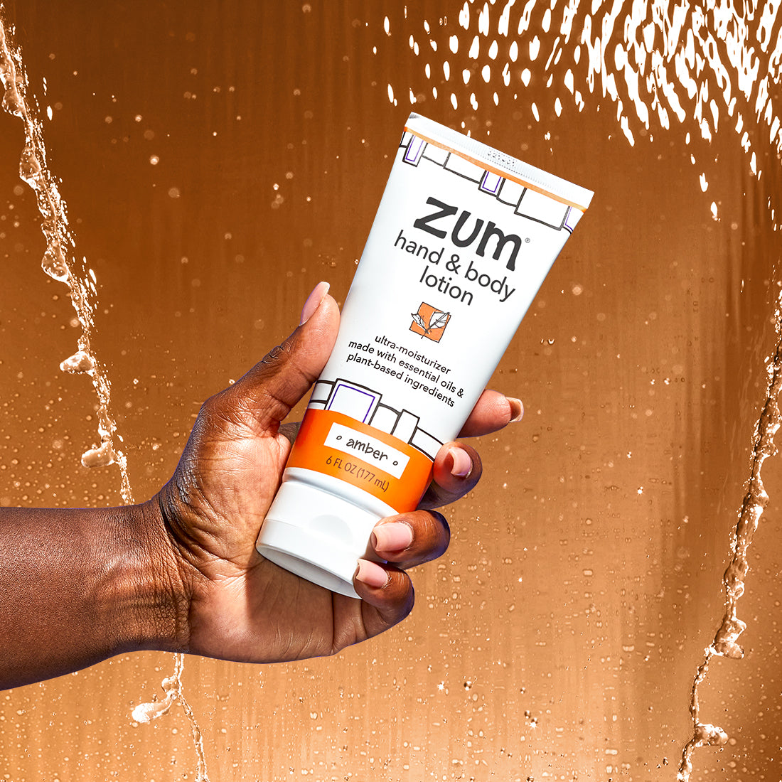 A hand holding from the left holding a 6 fl. oz. Amber hand & body lotion tube. Water falling behind the product with a orange-ish brown background color.