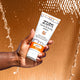 A hand holding from the left holding a 6 fl. oz. Amber hand & body lotion tube. Water falling behind the product with a orange-ish brown background color.