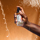 A hand coming from the right spraying an Amber mist. Water falling behind it and a brown background.