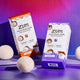 Tilted Amber and Lavender Wool Dryer Balls Kit boxes filled with three wool balls and an aroma blend, on a purple table, surrounded by three wool balls. Orange to purple gradient background.