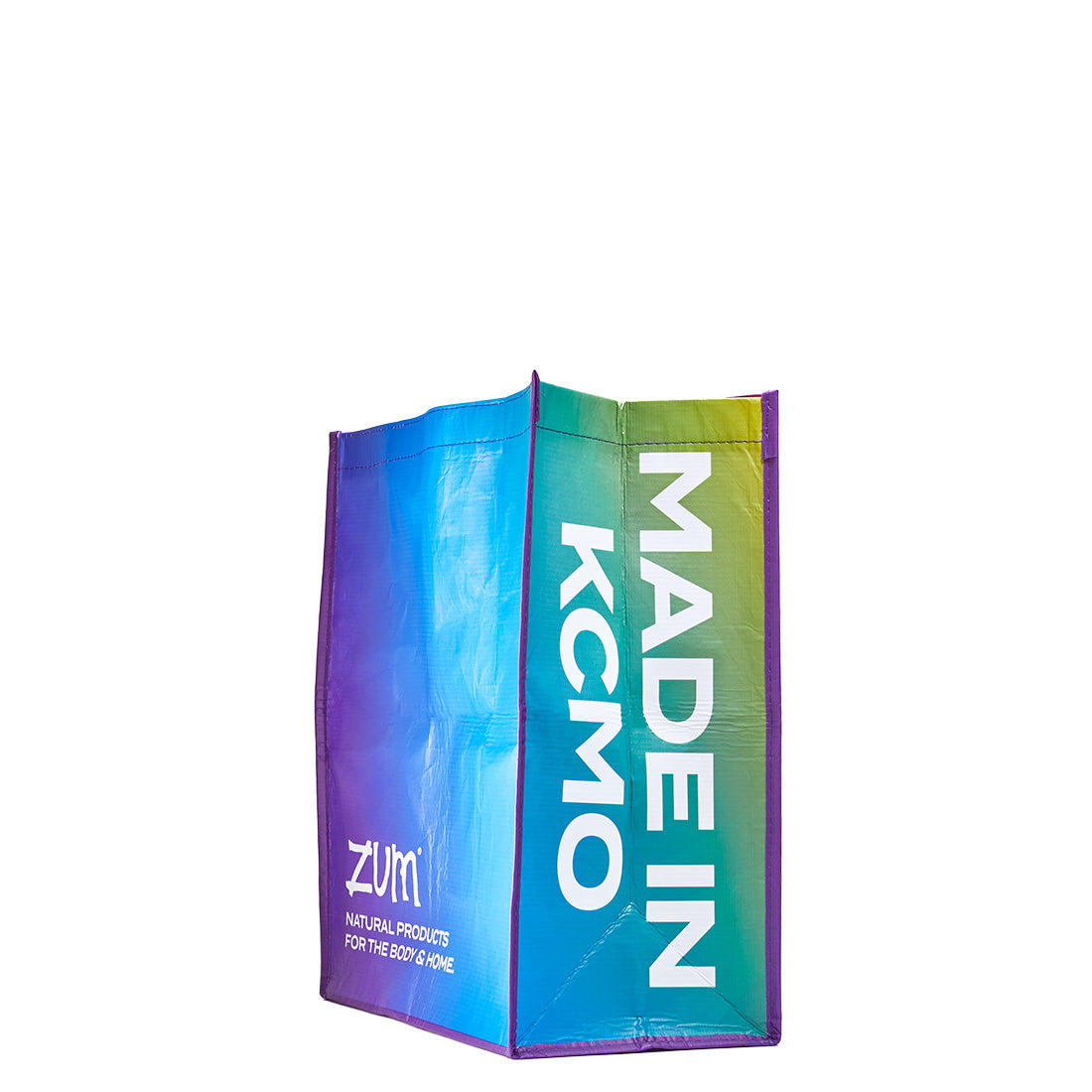 Side angle of recyclable tote bag. Made In KCMO phrase on the side of the bag, and Zum logo with phrase 'Natural products for the body and home' on the other side of the bag. Purple to blue to green gradient on the bag..