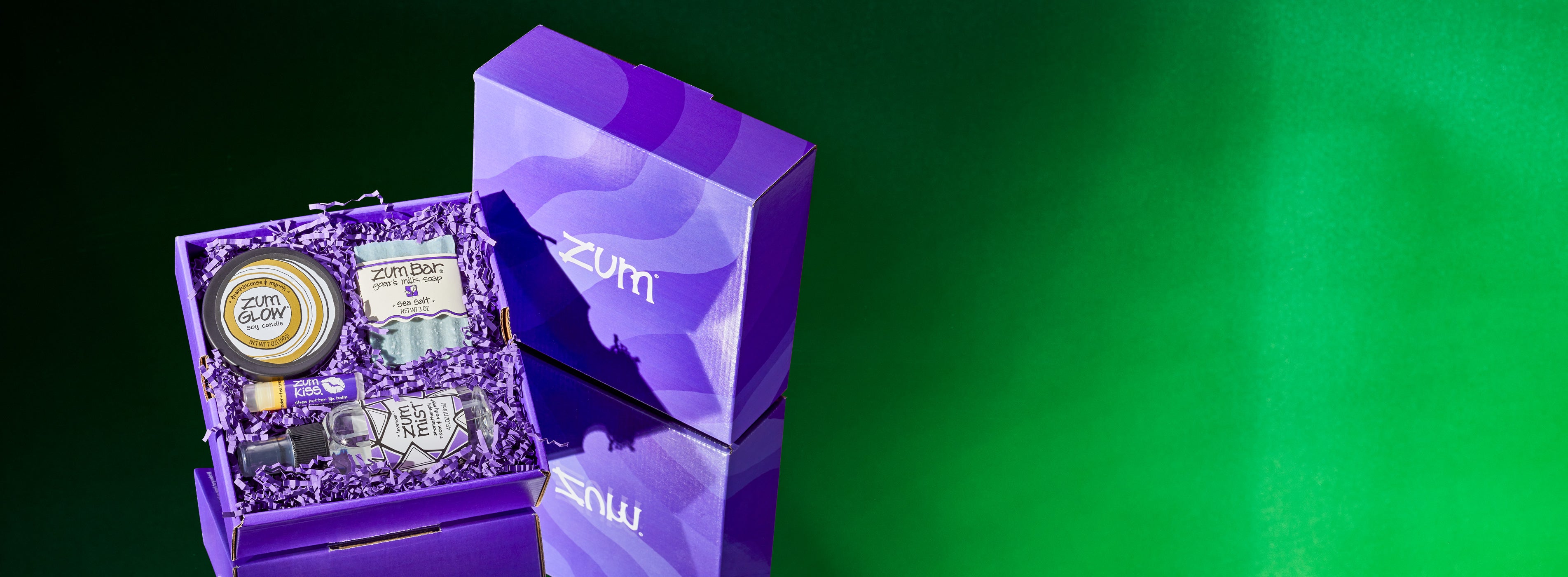 A tilted, open purple gift box with crinkle on a mirrored surface with a green gradient background. Outer box sitting next to it. On the crinkle there lays a Frankincense & Myrrh candle tin, Sea Salt bar soap, Tea Tree-Citrus lip balm, and Lavender mist bottle.