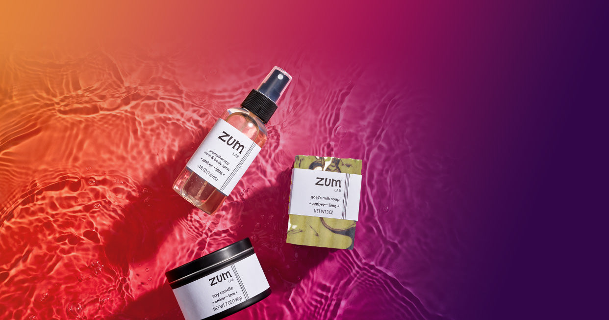Amber-Lime room & body mist bottle, goat's milk bar soap, and soy candle laying on a surface with water. Orange to purple gradient background.