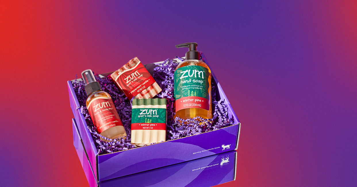 Purple box with purple crinkly laying on a mirrored surface with a purple and red gradient background. The box contains a Peppermint-Vanilla scented mist bottle and bar soap, and Winter Pine scented bar soap and hand soap bottle.