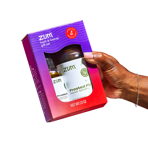 A hand coming from the right holding a Fresh & Festive Gift Set. The gift set consists of a purple to red gradient rectangular box with a Frosted Fir Mist and Hand Soap inside.