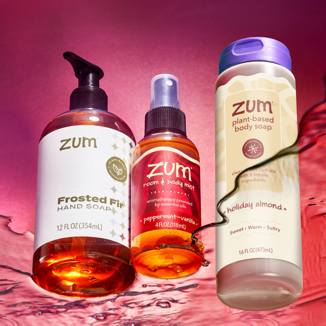 Three Zum holiday personal care products arranged together against a pink and red liquid splash background: a frosted hand soap bottle, a peppermint vanilla room and body mist spray bottle, and a holiday almond plant-based body soap bottle.