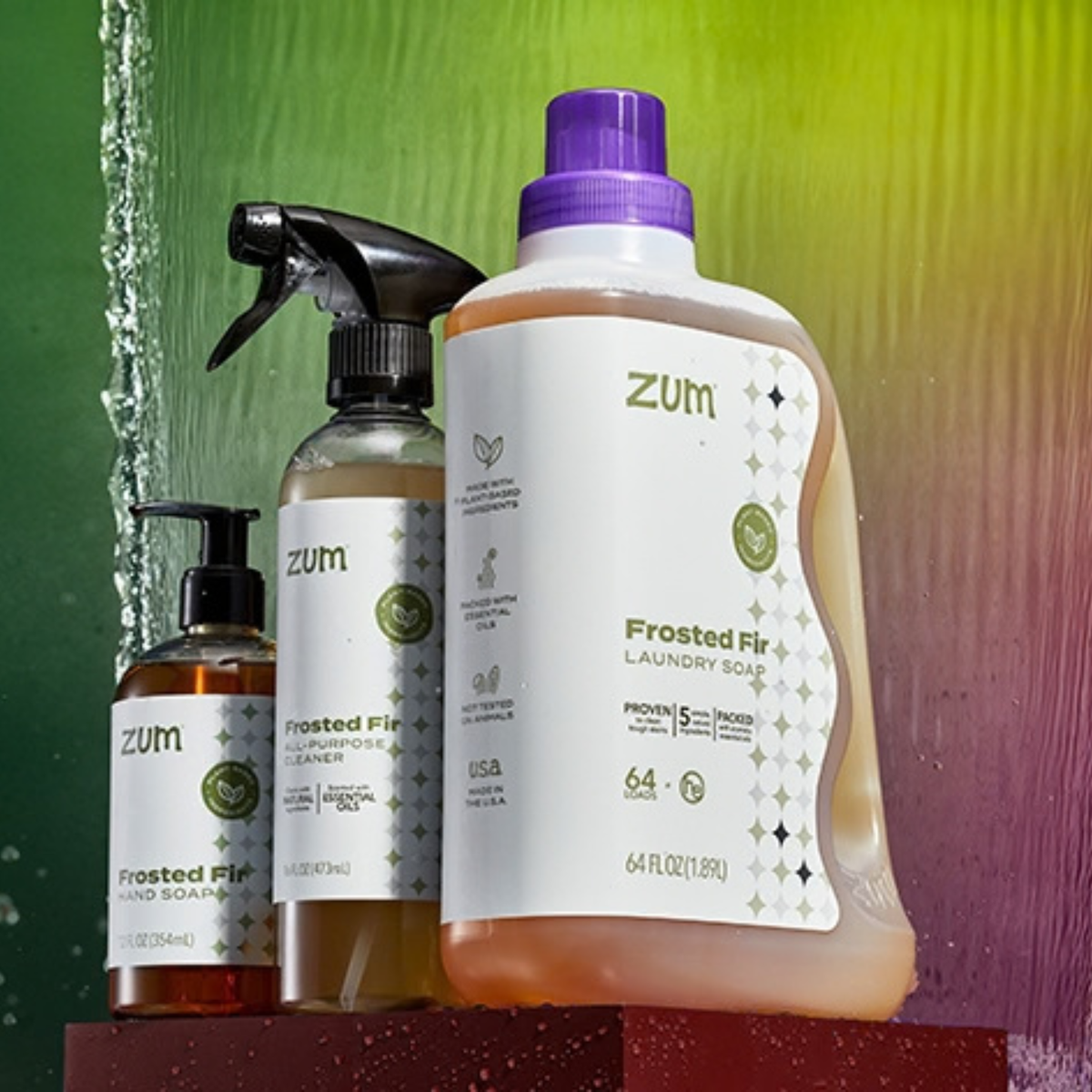 Three Zum frosted fir home care products displayed together: laundry soap bottle with a purple cap, a frosted all-purpose cleaner spray bottle, and a hand soap bottle, set against a green-to-purple gradient background with water droplets.