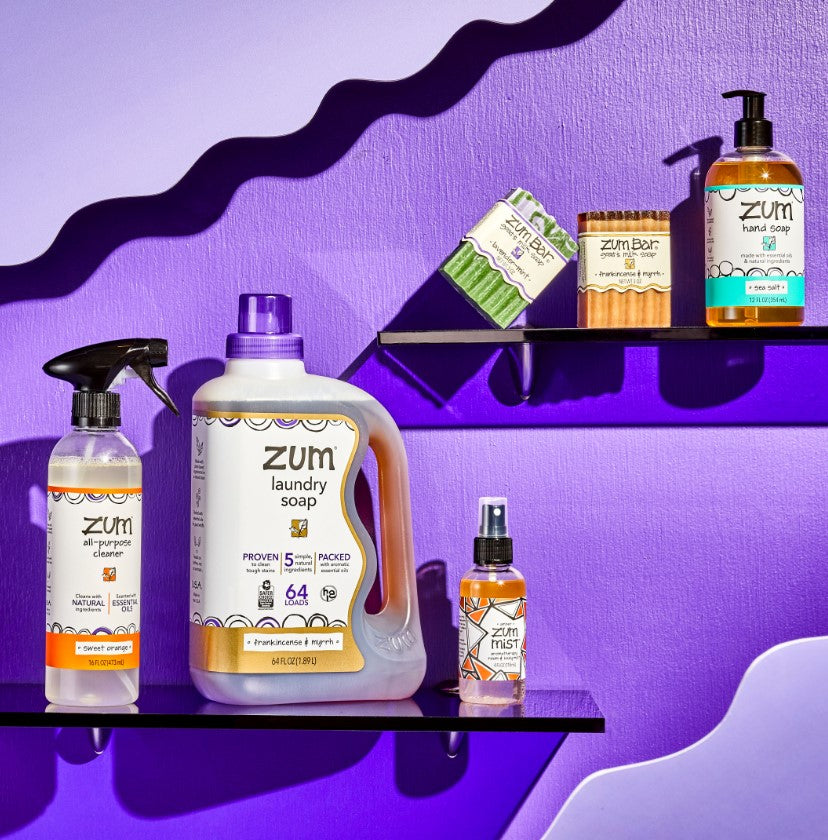 Sweet Orange All-Purpose Cleaner, Frankincense & Myrrh 64 oz Laundry Soap and Zum Bar, Lavender-Mint Zum Bar, Sea Salt Hand Soap, and Amber Mist on two purple shelves.