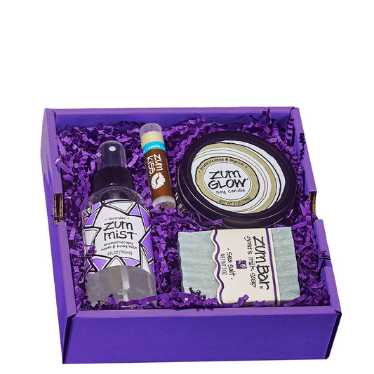 Square gift box containing Mist, lip balm, bar soap and candle in purple crinkle paper.